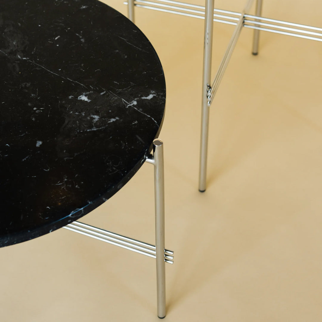 Round black marble table with white veining, supported by sleek silver metal legs on a beige background.