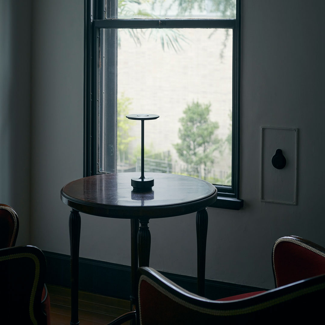 Black metal stand on a dark wood table, framed by neutral-toned walls and greenery outside the window.