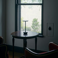 Black metal stand on a dark wood table, framed by neutral-toned walls and greenery outside the window.