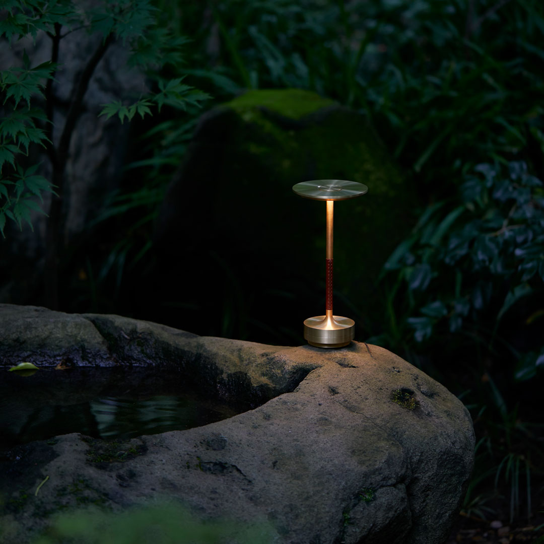 Sleek metallic gold lamp with a glass shade, set against rich green foliage and dark rock textures.