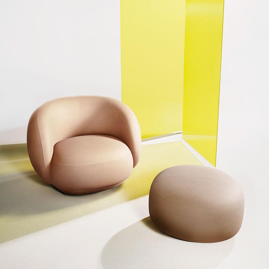 Curved beige armchair and round ottoman with a vibrant yellow backdrop, showcasing smooth, modern design and soft tones.