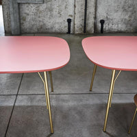 Two pink tables with smooth surfaces and sleek gold legs set against a concrete backdrop. Stylish contemporary design.