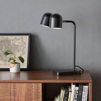 Stylish black desk lamp with dual shades, set against a gray wall and warm wood surface, complemented by greenery.