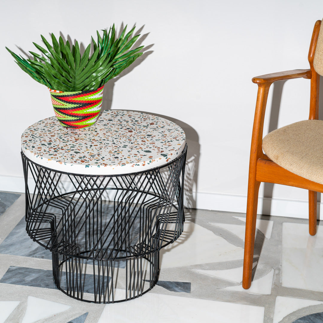Round table with a colorful speckled top and black geometric wire base, complemented by a vibrant striped planter.