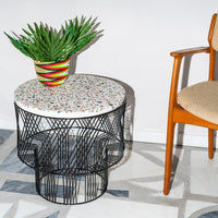 Round table with a colorful speckled top and black geometric wire base, complemented by a vibrant striped planter.