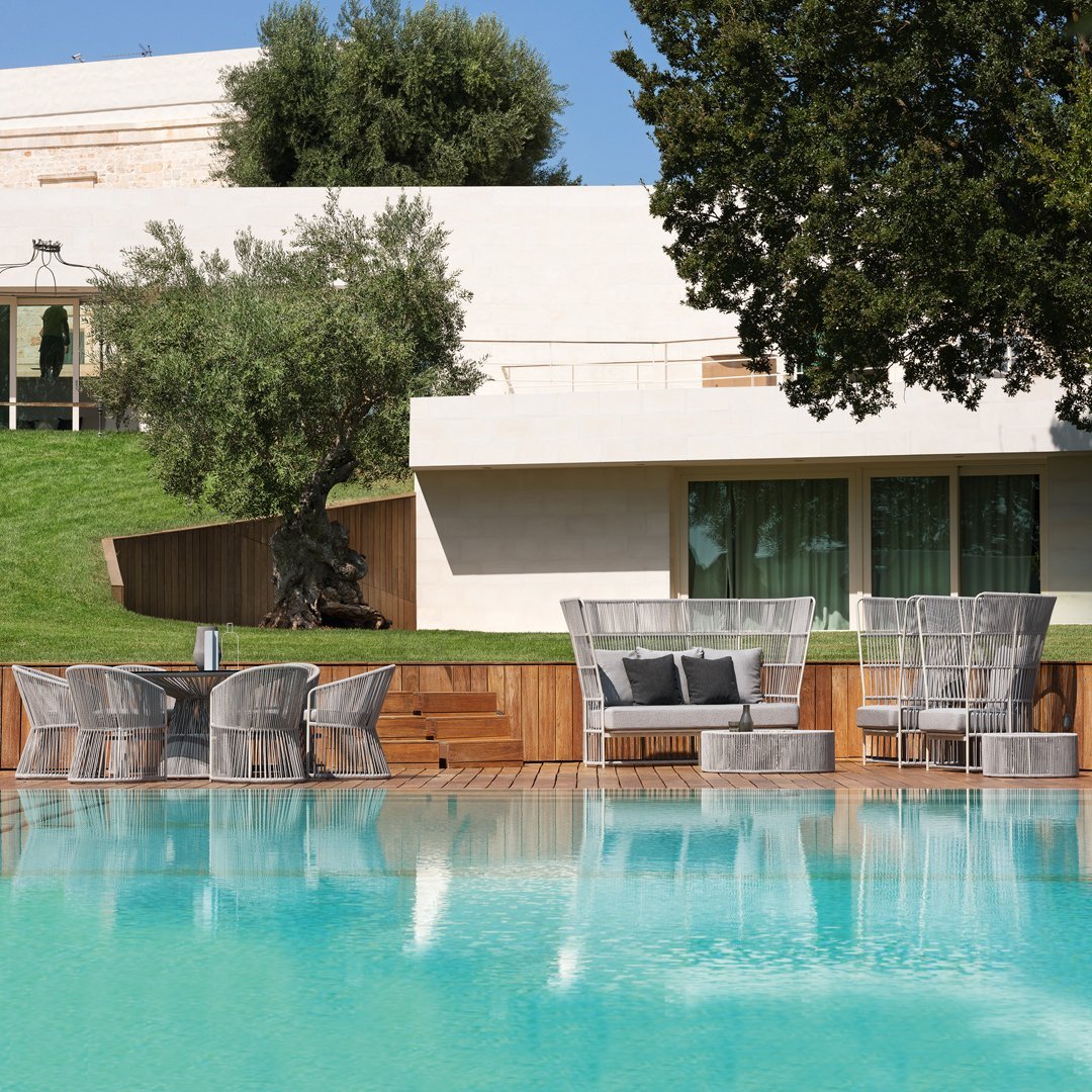 Elegant outdoor furniture set with gray wicker pattern, surrounded by lush greenery and a tranquil blue pool.