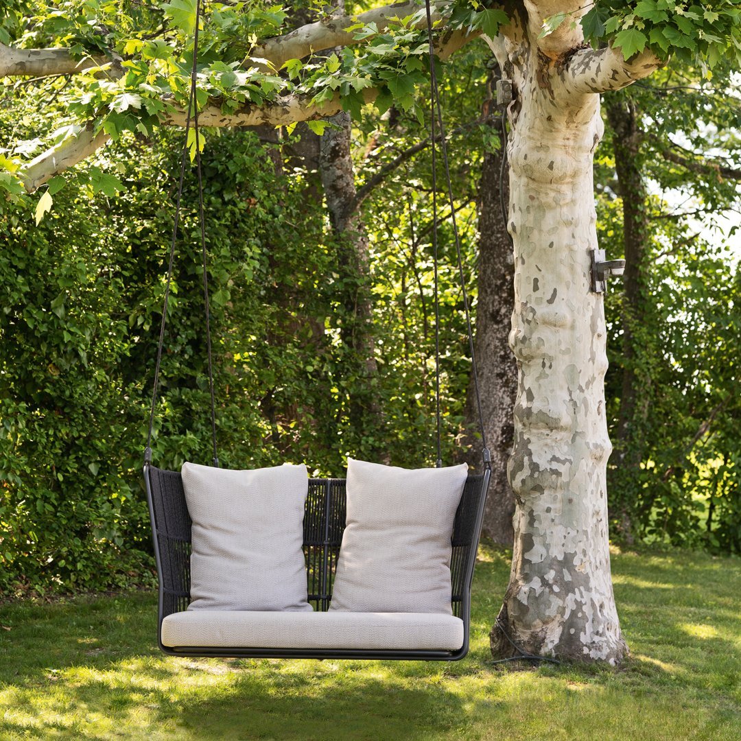 Stylish black wicker swing with beige cushions, set against lush green foliage and a tree backdrop.
