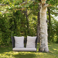 Stylish black wicker swing with beige cushions, set against lush green foliage and a tree backdrop.