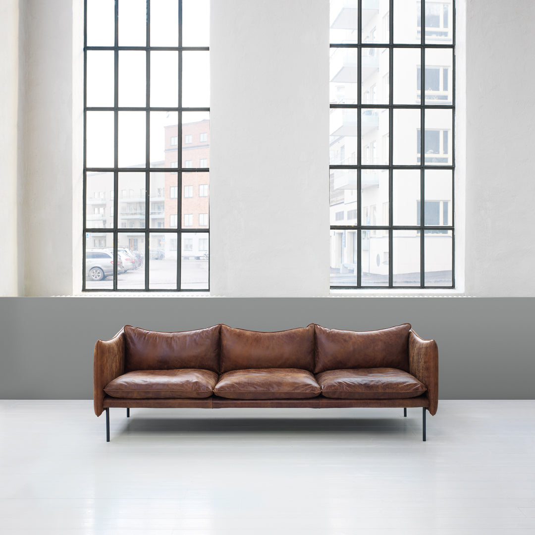 Brown leather sofa with a sleek design, featuring smooth textures against a minimalistic gray wall backdrop.