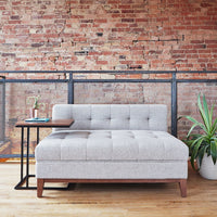 Light gray tufted upholstery couch with mid-century wood legs, set against a rustic brick wall backdrop.