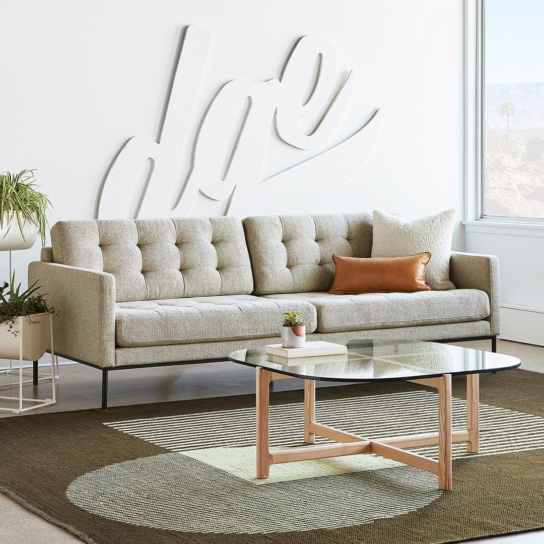 Beige tufted sofa with colorful accent pillow on a patterned rug featuring green and brown circular designs under a clear table.