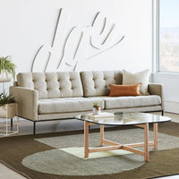 Beige tufted sofa with colorful accent pillow on a patterned rug featuring green and brown circular designs under a clear table.