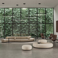 Modern beige sofa and round ottoman on a concrete floor, with lush greenery visible through large windows.