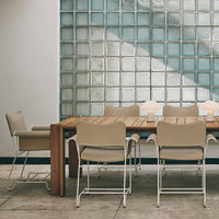 Beige outdoor dining chairs paired with a wooden table, set against a textured glass block wall. Modern, neutral tones.