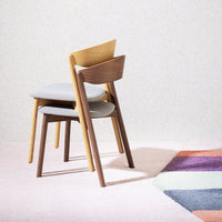 "Stacked chairs in warm wood tones on a soft pink floor with a colorful geometric rug design."