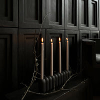 Neutral beige candles atop a sleek black candle holder, set against a dark backdrop with textured paneling.