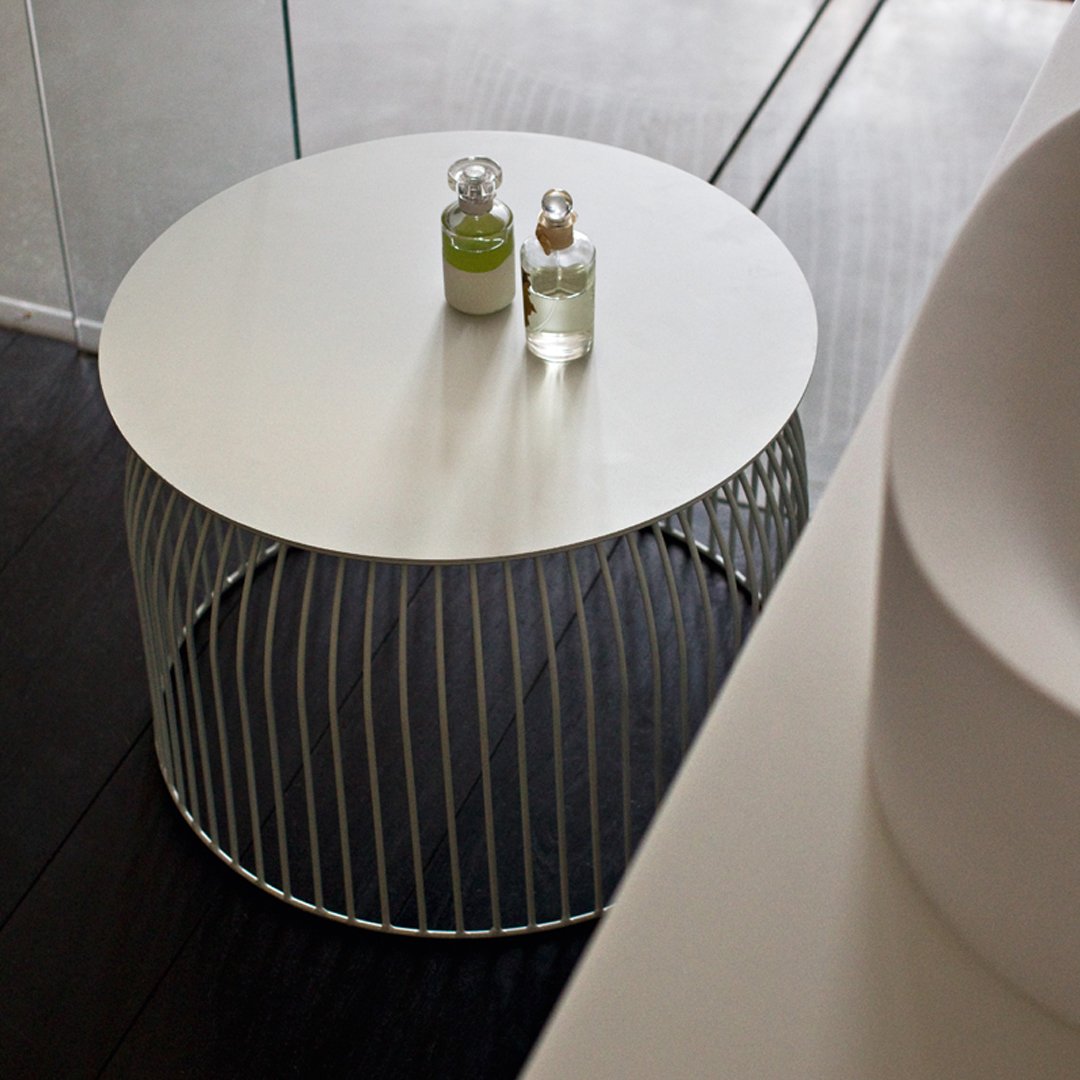 Round white tabletop with a linear wire metal base, set against dark wooden flooring. Elegant minimalistic design.