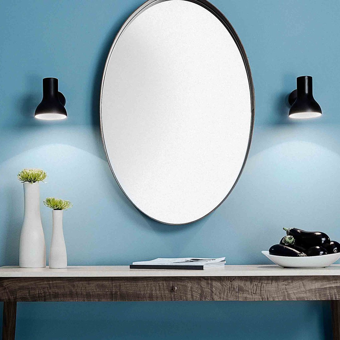 Modern blue wall with a round mirror, black wall sconces, and a wooden table featuring white vases and a black plate.