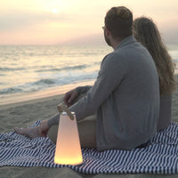 "Soft glow lamp beside couple on striped blue and white blanket at sunset on the beach."