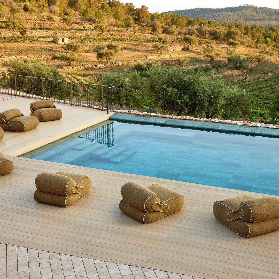 "Neutral brown roll cushions arranged around a clear blue pool with natural green hills in the background."