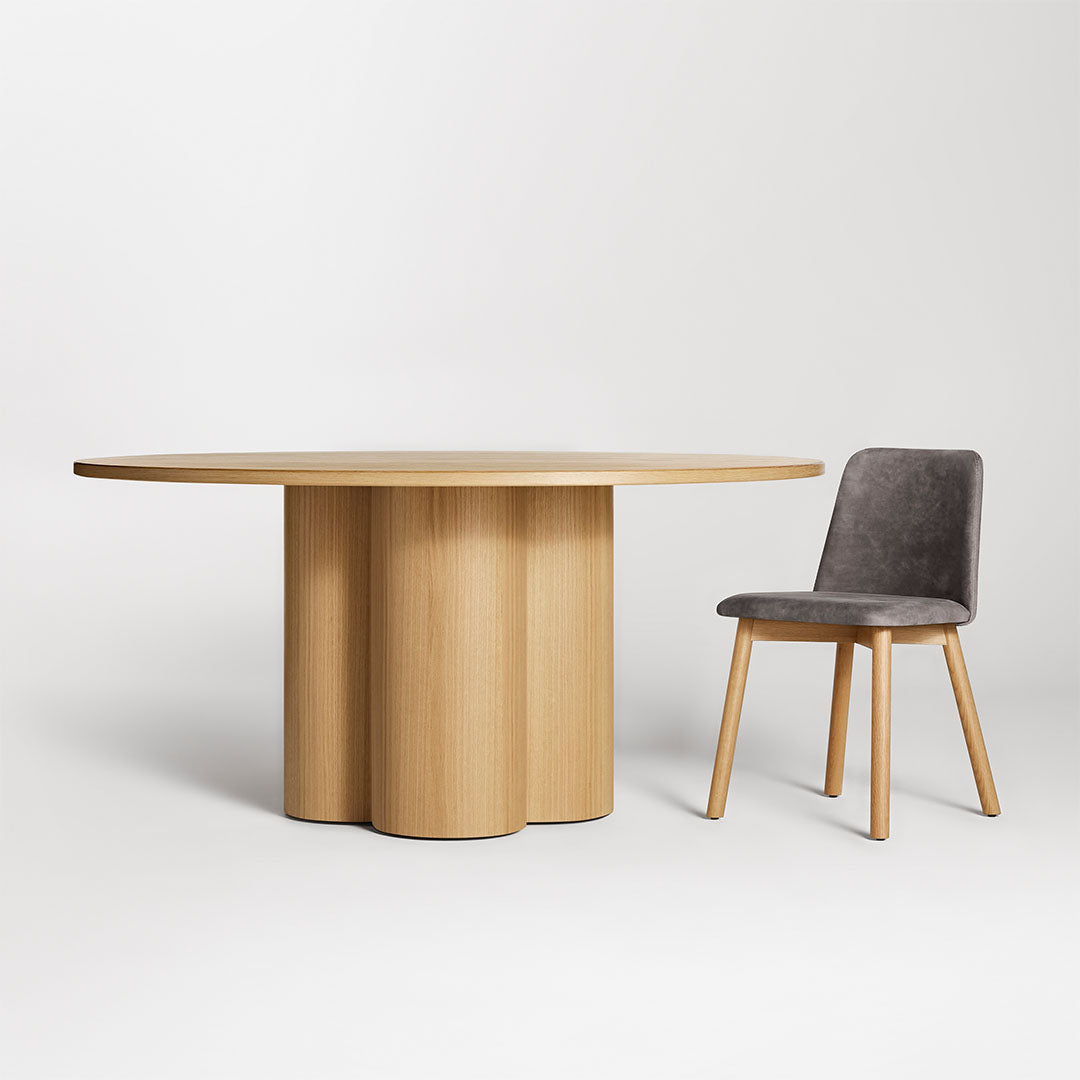 Natural wood round table with a scalloped base and a grey velvet chair, creating a modern and minimalist look.