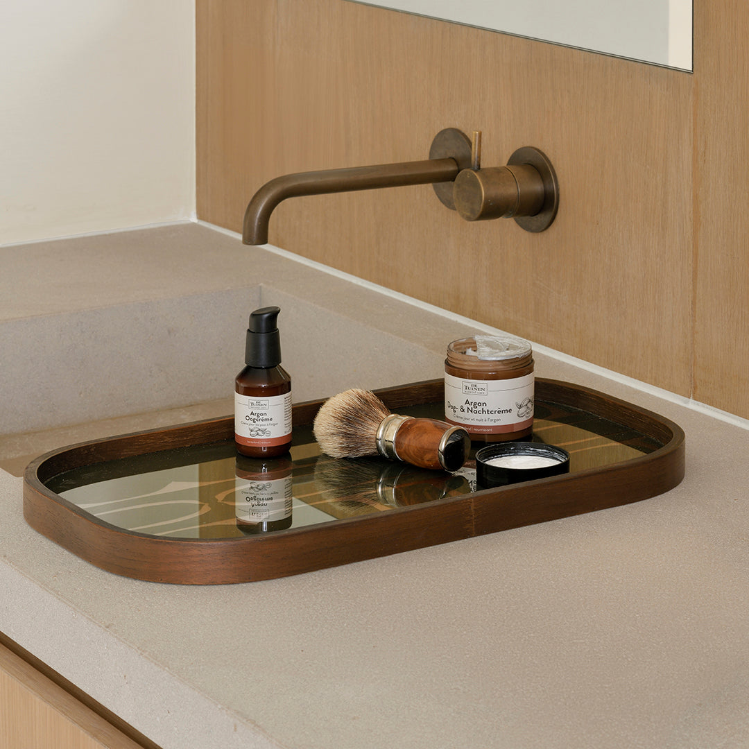 "Bronze oval tray with mirrored surface, holding black and brown grooming products on a beige countertop."