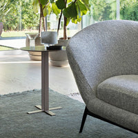 Gray textured fabric armchair and sleek metallic side table on a light floor with green plants in the background.