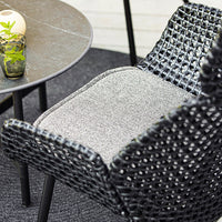 Black woven chair with a gray cushion, featuring a textured pattern, paired with a round black table and greenery.