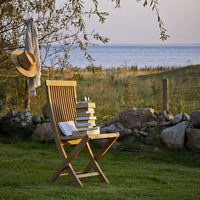 "Light wooden folding chair with a natural finish, surrounded by green grass and stone, with soft pastel sky in the background."