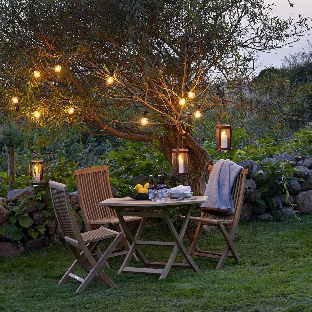 Wooden chairs and round table, lit by warm glowing lanterns, set against lush green grass and a stone wall.