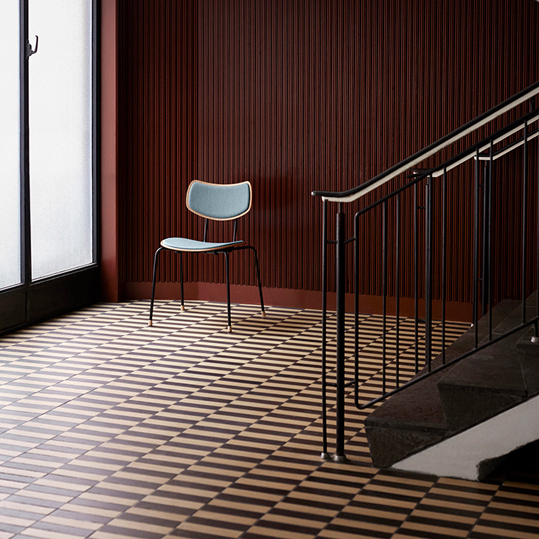 Light blue chair on patterned black and cream tile floor against a deep red wooden wall. Modern, minimalist design.