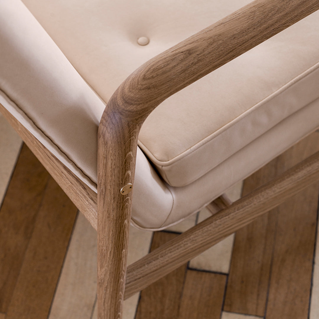 Beige cushioned chair with natural wood frame and subtle button detail, set against a patterned wooden floor.