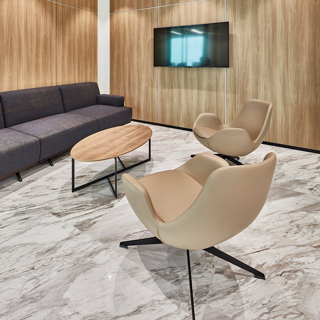 Modern lounge with dark gray sofa, beige chairs, and polished marble flooring, complemented by wood panel walls.