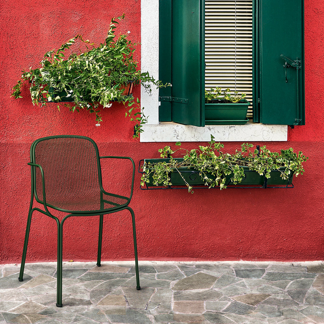 Dark green metal chair against a vibrant red wall with green shutters and lush plants, creating a bold, colorful contrast.