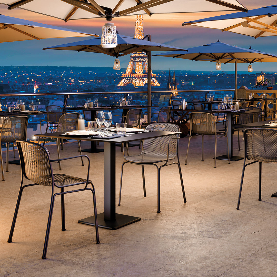 Elegant grey metal chairs and tables under beige umbrellas, set against a twilight skyline with the Eiffel Tower.