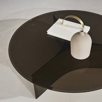 Dark brown circular glass table with subtle geometric shadows, styled with a white book and a minimalist gray vase.