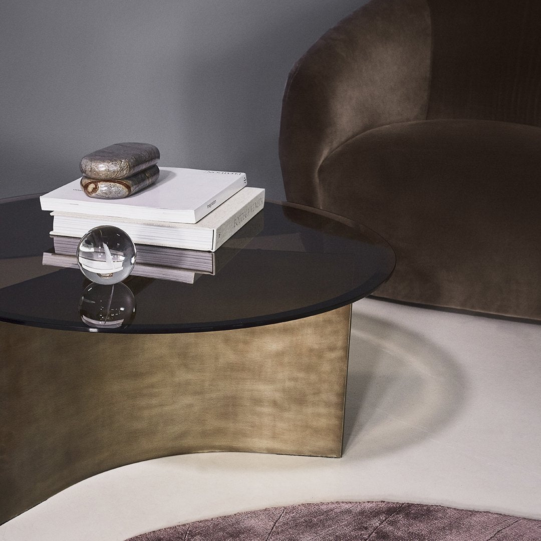 "Round black glass tabletop with a brushed gold base, surrounded by a plush brown chair and soft pink rug."