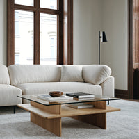 Beige upholstered sofa with soft curves, paired with a light wood coffee table and glass top, set against neutral tones.