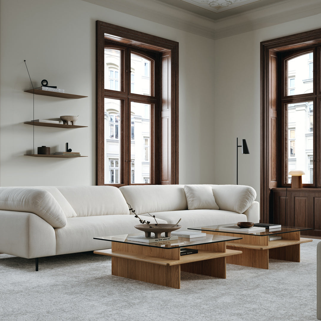 Modern beige sofa with textured upholstery, wooden coffee tables, and neutral gray carpet in a stylish, light-filled room.