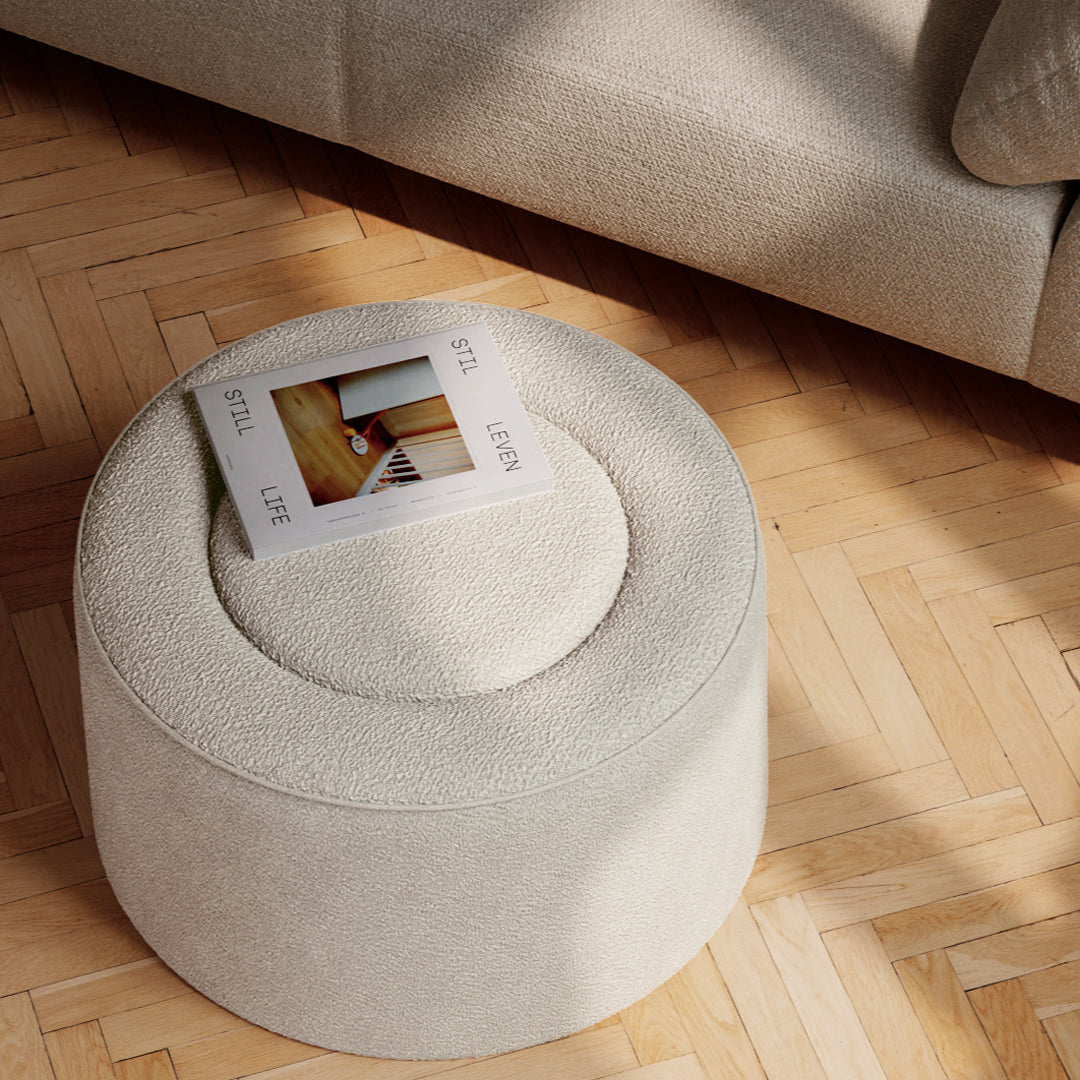 Round cream ottoman with textured fabric on a wooden herringbone floor, featuring a circular top and seamless design.