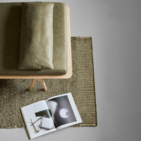 Olive green cushion, textured beige rug, and minimalist magazine on a light gray background.
