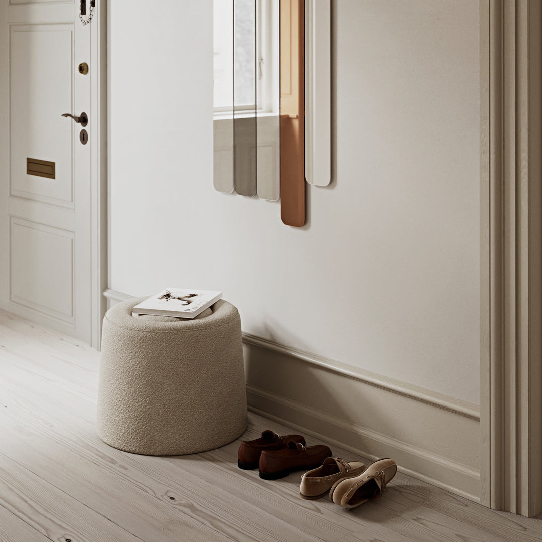 Neutral beige pouf with smooth texture beside brown and cream shoes, set against a light wooden floor and soft wall colors.