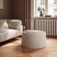 Beige round ottoman with a textured pattern, set on a warm wooden herringbone floor in a cozy, neutral-toned room.