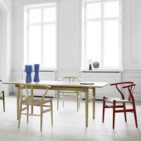 Light wood dining table with blue, green, and red chairs, set in a bright, airy white room. Modern minimalist design.