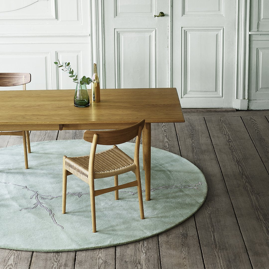 Wooden dining table with natural finish, surrounded by a beige chair on a soft, round green marble-patterned rug.