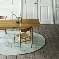 Wooden dining table with natural finish, surrounded by a beige chair on a soft, round green marble-patterned rug.