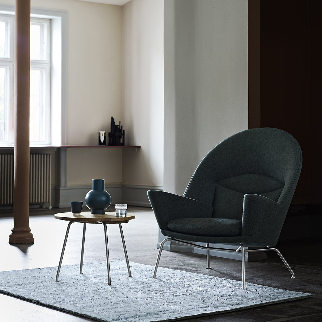 Dark teal chair on a light gray rug, featuring a minimalist wooden table and subtle lighting. Ideal for modern decor.