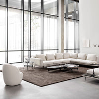 Modern white sectional sofa on a dark brown rug in a bright, minimalist living room with large windows.