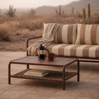 Brown metal coffee table with woven shelf, paired with striped beige and cream cushions. Perfect for outdoor decor.