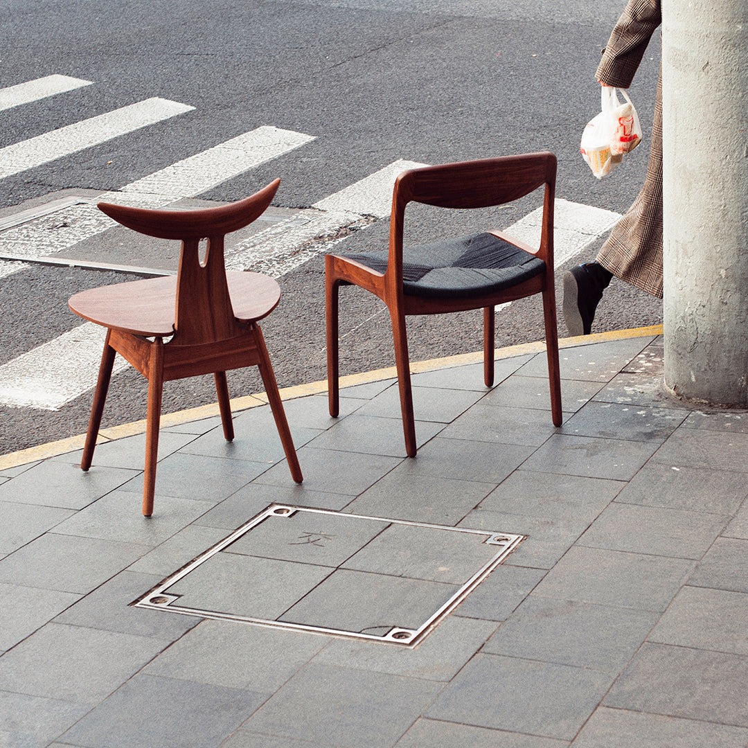 "Pair of modern wooden chairs in rich brown tones with sleek black seating, placed on a textured gray pavement."